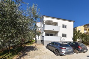 A two-story building with a white facade, a balcony on the second floor, and two cars parked in front.