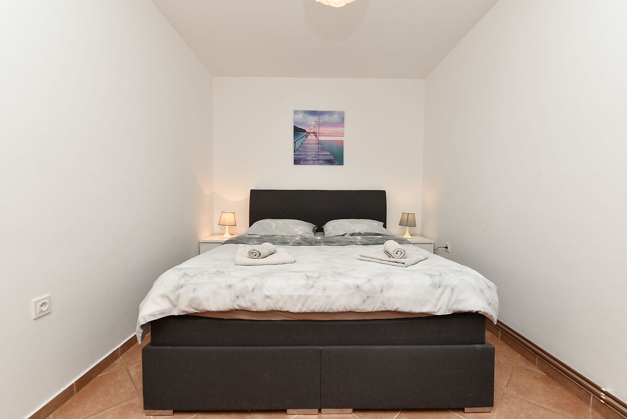 A bedroom with a bed, two bedside lamps, a wall-mounted picture, and a small bedside table.