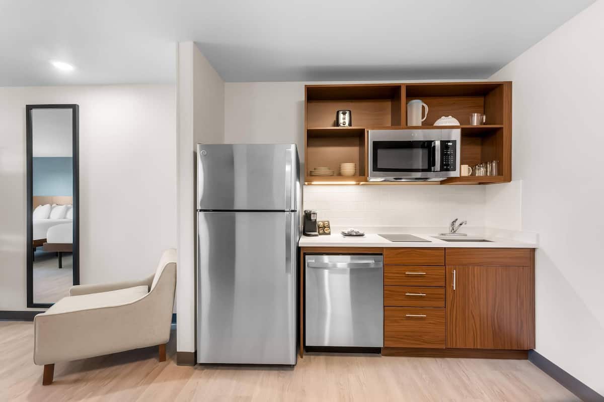 A modern kitchen with a stainless steel refrigerator, microwave, and dishwasher, wooden cabinets, and a built-in shelf with kitchenware.