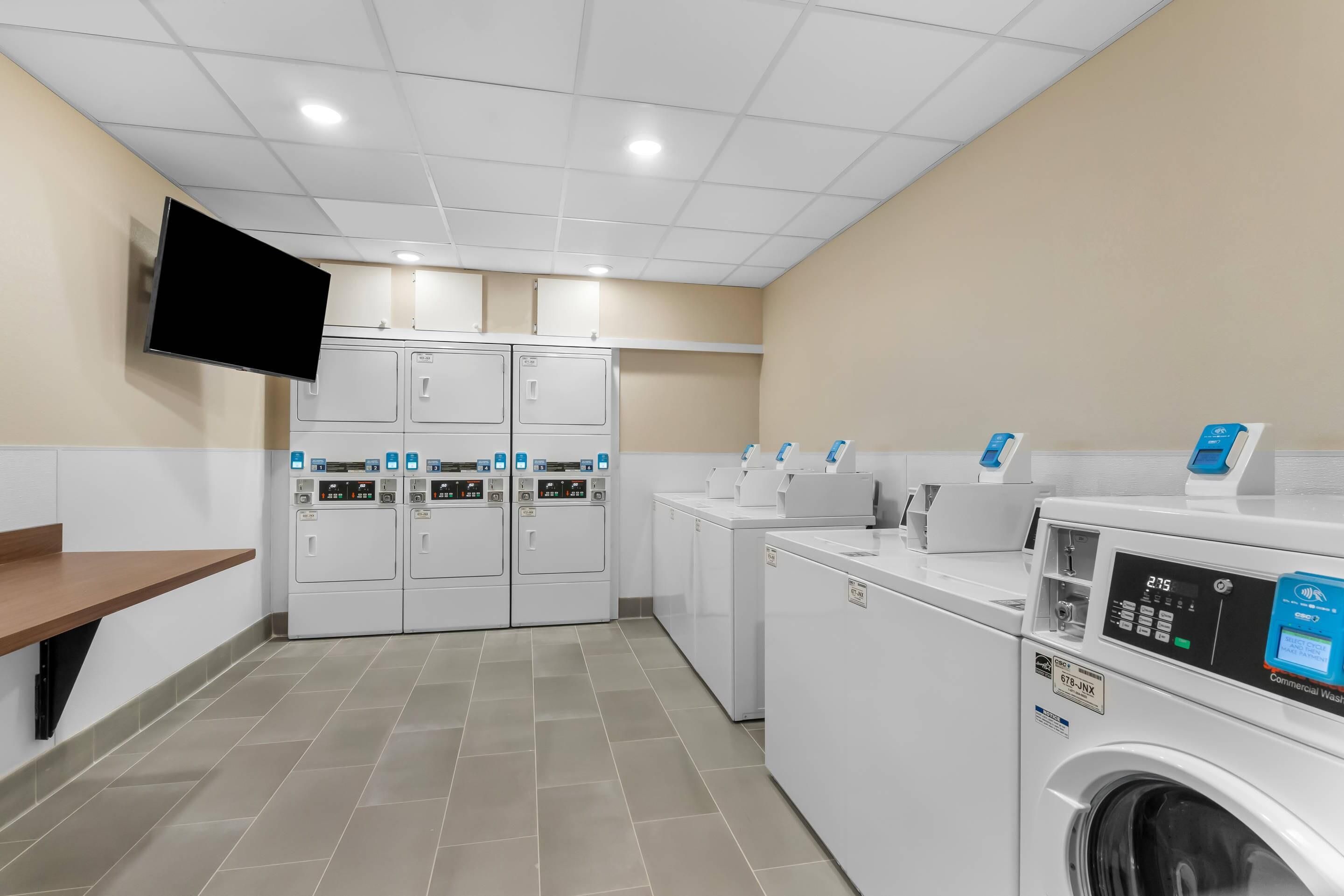 Une buanderie équipée de plusieurs machines à laver et de plusieurs sèche-linge, d’un téléviseur à écran plat fixé au mur et d’un sol carrelé.