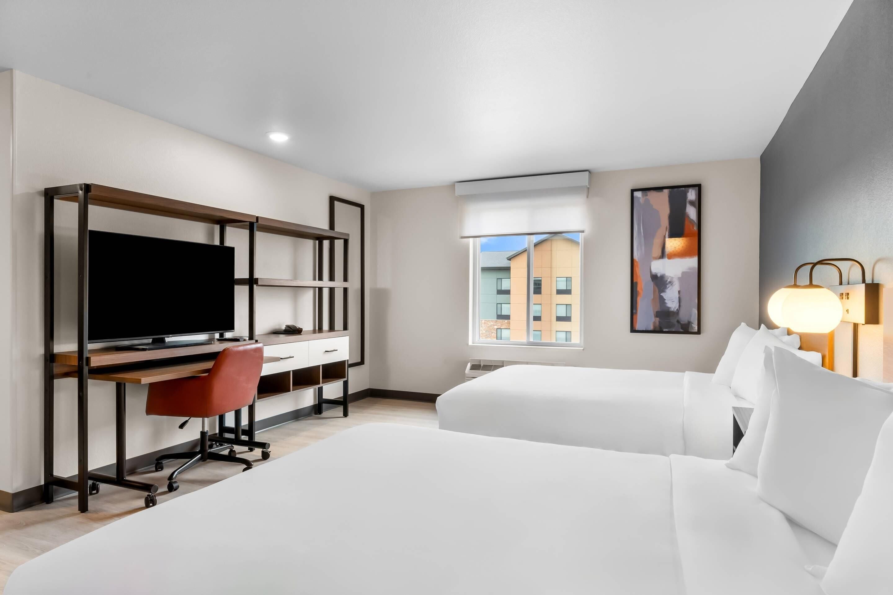 Habitación de hotel con una cama grande, un escritorio con televisor, una silla, una ventana con vista a edificios y una lámpara.