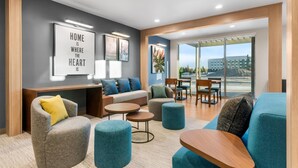 Un salon moderne comprenant un canapé, des fauteuils, des poufs et un coin repas avec une table et des chaises. Des tableaux encadrés ornent le mur, et une grande fenêtre offre une vue sur l’extérieur.