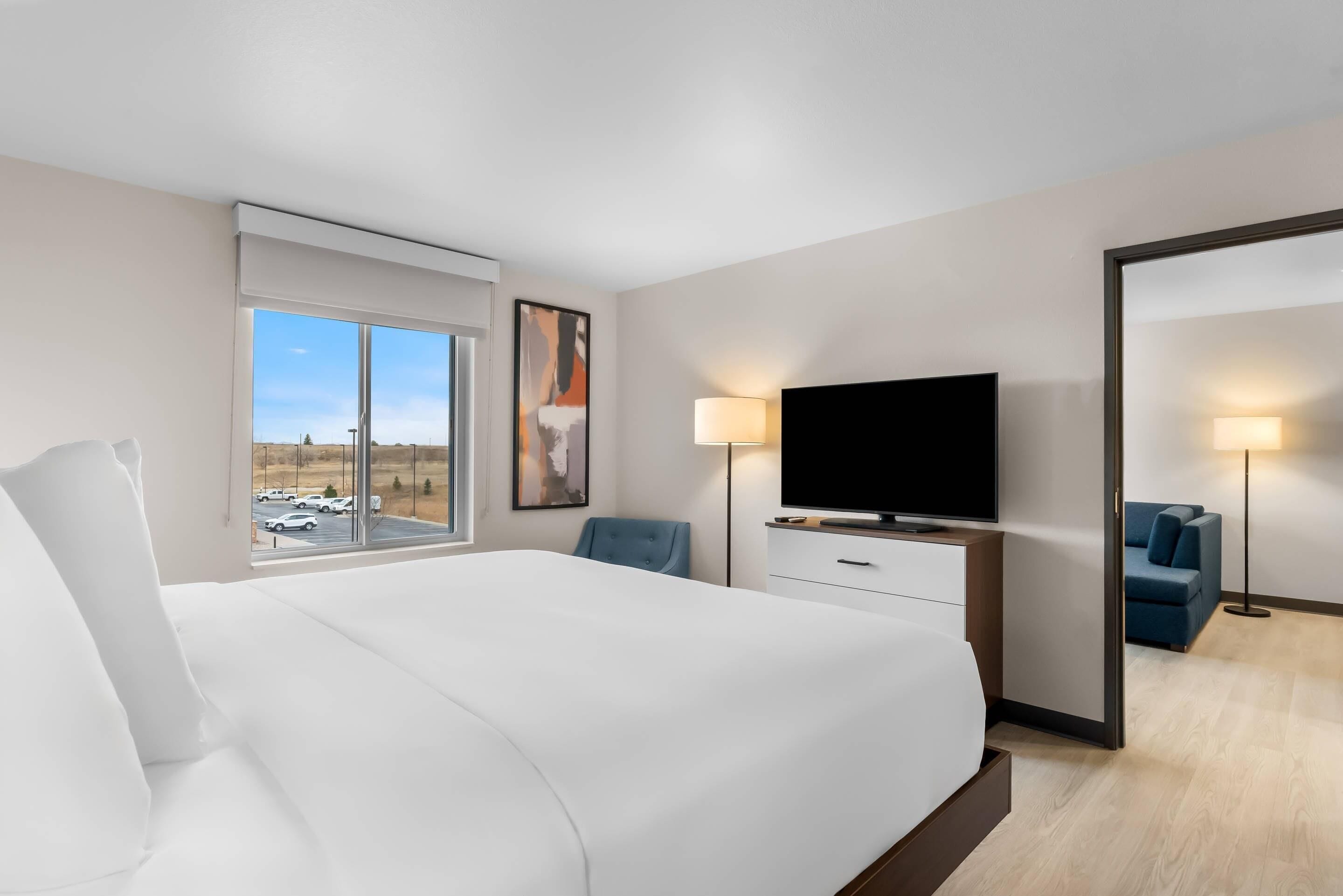A hotel room with a large bed, a flat-screen TV, a desk with a lamp, a chair, and a large window with a view of parked cars and a building.
