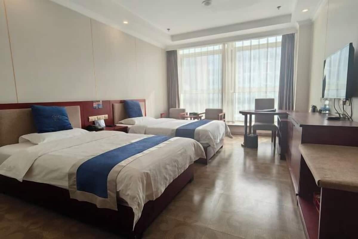 Ein Hotelzimmer mit zwei Betten, einem Schreibtisch, einem Stuhl, einem Fernseher und einem großen Fenster mit Vorhängen.