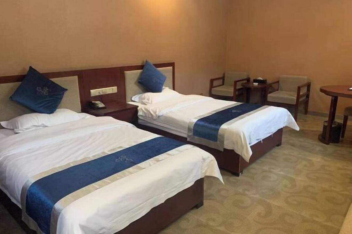 Ein Hotelzimmer mit zwei Betten, beide mit blauer Bettdecke und weißen Laken. Es gibt zwei Nachttische mit Lampen, einen kleinen Tisch und zwei Stühle.
