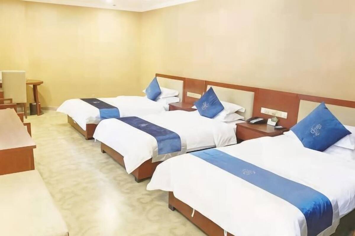 Ein Hotelzimmer mit drei Betten, jeweils mit einer blau-weißen Tagesdecke, einem Holz-Kopfteil und einem Nachttisch mit Telefon.