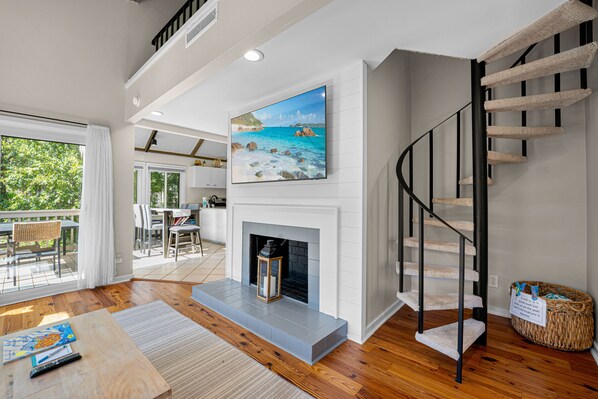 A spacious living area with a fireplace, a staircase with a cat tree, and a large window overlooking greenery.