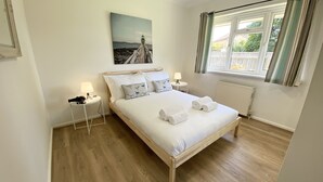 A neatly made bed with white linens and rolled towels, a bedside table with a lamp, and a painting of a lighthouse on the wall. - Daisy Cottage by NewFo rest (Lymington)