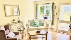 A cozy living room with a sofa, armchair, and coffee table. A dresser with a mirror and lamp is against the wall. A window with curtains offers a view of greenery outside. - Daisy Cottage by NewFo rest (Lymington)
