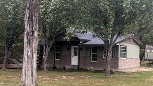Une petite maison de plain-pied, avec une façade en briques et une porte blanche, entourée d’arbres et d’une pelouse.