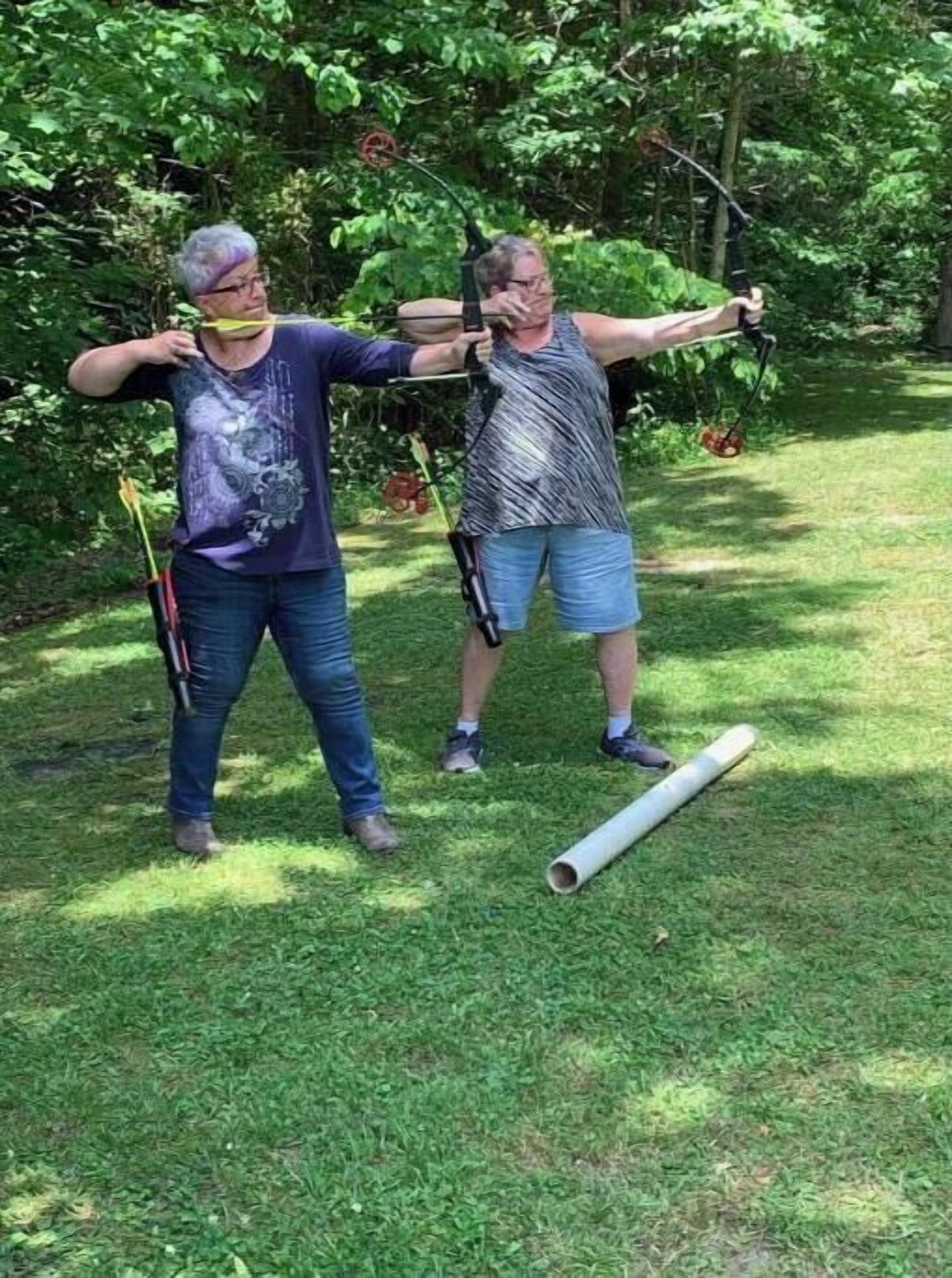 Two people practicing archery in a grassy outdoor area with trees in the background.