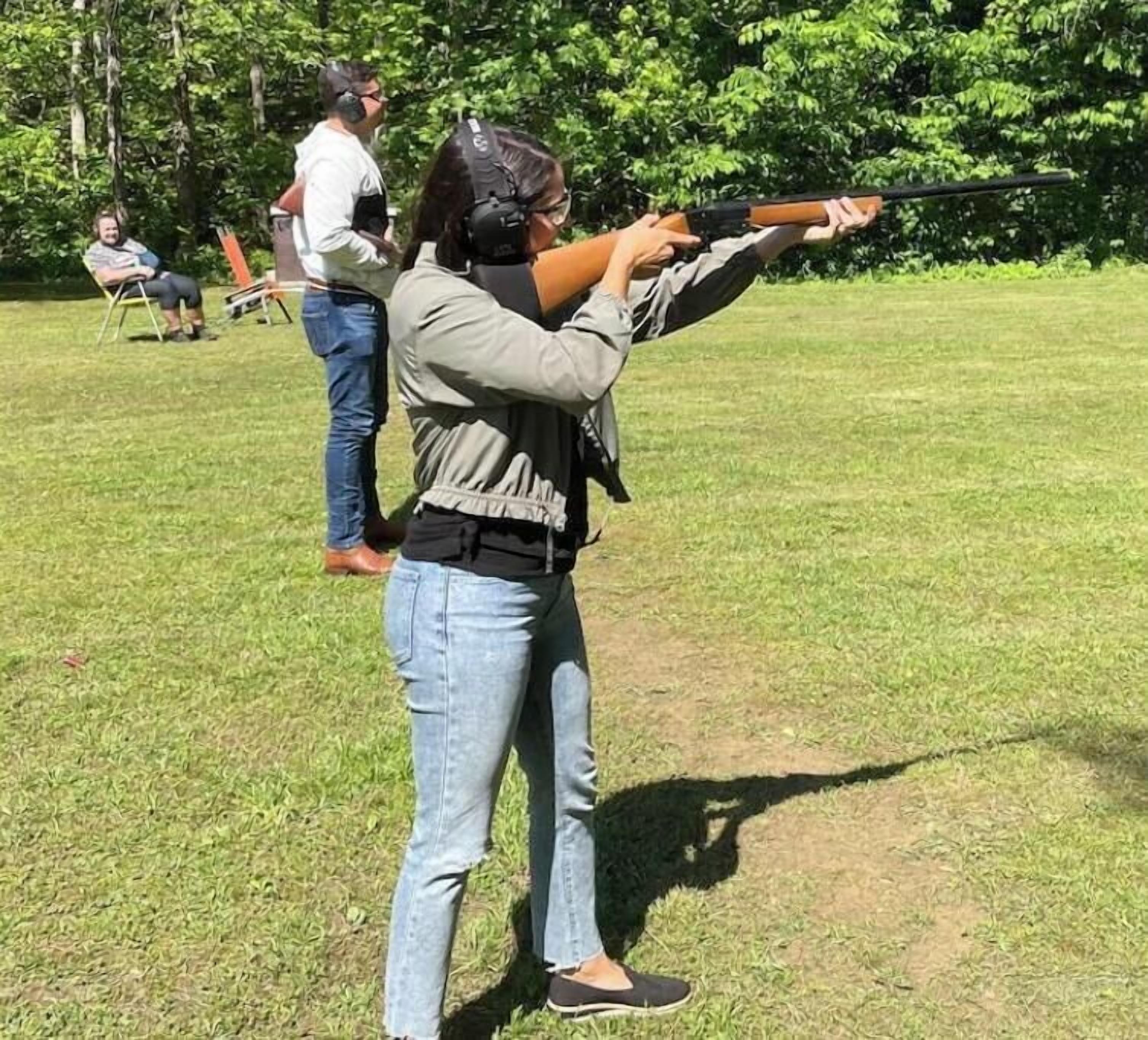 A person aiming a rifle in an open field with grass and trees in the background.