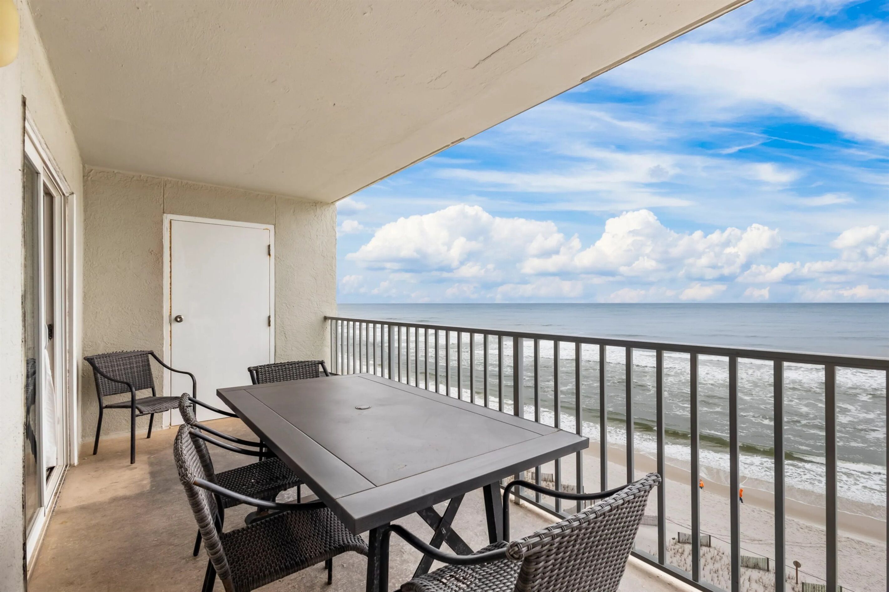 A balcony with a table and chairs, overlooking the ocean with a view of the beach and clouds.