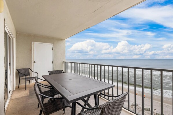A balcony with a table and chairs, overlooking the ocean with a view of the beach and clouds.