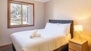 Ein ordentlich bezogenes Bett mit weißen Leinen und einem aufgerollten Handtuch, eine Nachttischlampe und ein Fenster mit Blick auf Bäume und einen Zaun.
