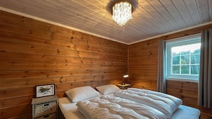 A cozy wooden room with a bed, a nightstand, a lamp, and a window with a view of trees.