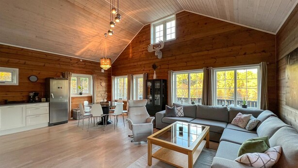 A spacious cabin interior with a dining area, sofa, and a large window offering a view of autumn foliage.