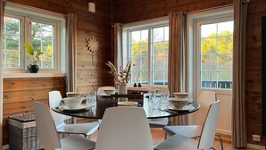 A dining area with a round table, white chairs, and a view of autumn trees through large windows.