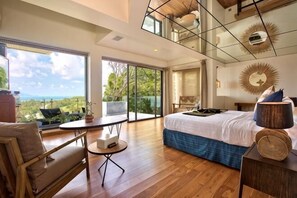 Quarto com cama grande, mesa redonda, sofá e vista para o exterior.