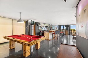 Una mesa de billar en una habitación moderna con zona de bar y vistas al exterior.
