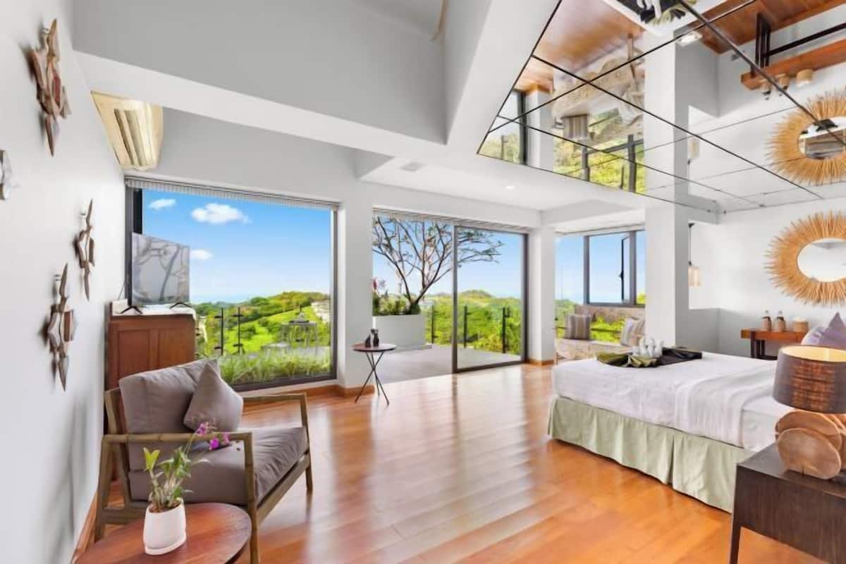 A spacious bedroom with a large bed, a seating area with a chair and table, and a balcony with a view of the countryside.
