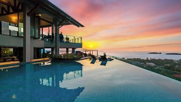 A modern infinity pool with a sunset view, a covered outdoor seating area, and a large building with multiple balconies. - Villa Skyfall : Spectacular 8 Bedroom Pool Villa with Private Cinema (Koh Samui)