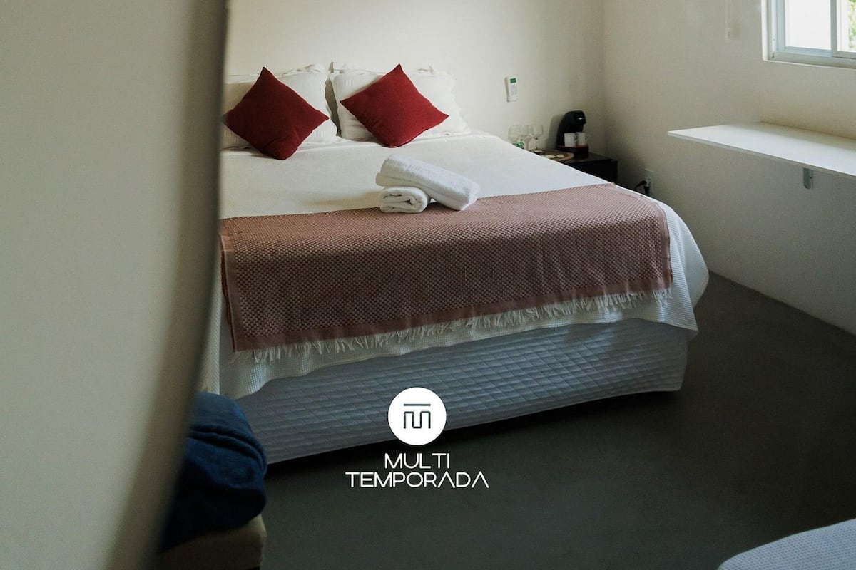 A bedroom with a bed, pillows, a hanging light fixture, a window, and a wall-mounted air conditioner.