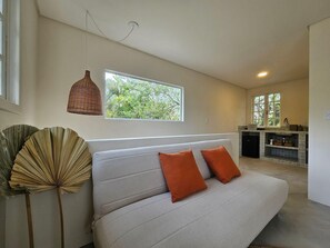 Ein modernes Wohnzimmer mit einer Couch, einem Fenster mit Blick auf Grünflächen, einer Pendelleuchte und einer Küche im Hintergrund.