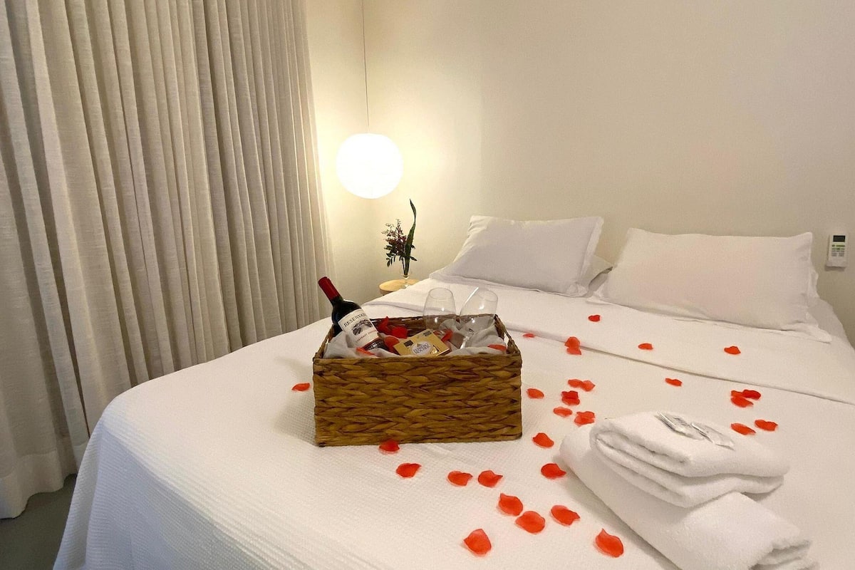 A hotel room with a neatly made bed, a bedside table with a bottle and glass, and rose petals scattered on the bed.