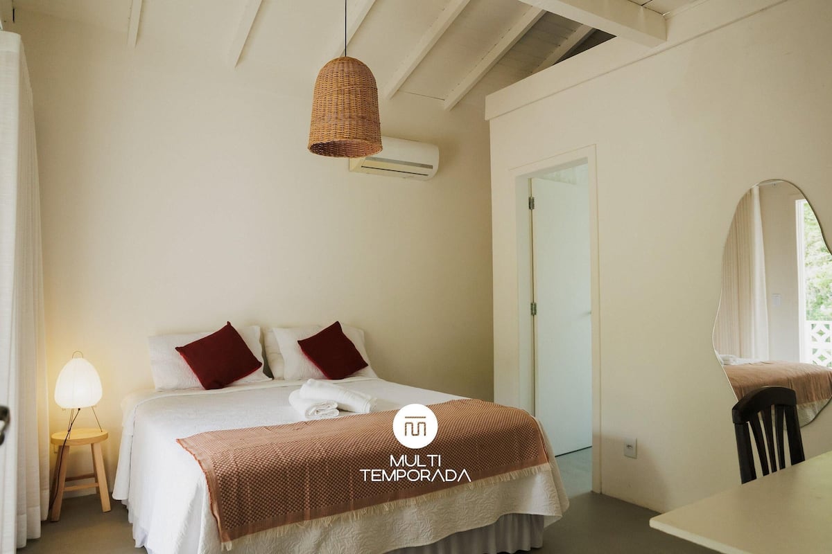 A hotel room with a bed, a bedside lamp, a hanging light fixture, a mirror, and an air conditioning unit.