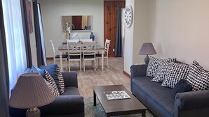 Un salon moderne avec un canapé, une table basse et une lampe. On aperçoit à l’arrière-plan un coin repas avec une table et des chaises.