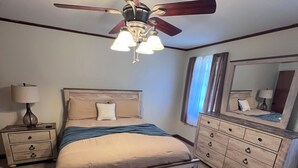 Une chambre à coucher comprenant un lit, un banc, une commode, une table de chevet, une lampe, un ventilateur de plafond et un miroir.