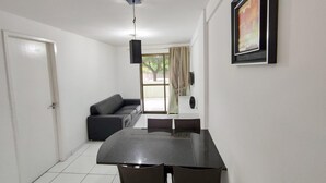 A modern living room with a sofa, a dining table with chairs, a TV mounted on the wall, and a door leading to another room. - Cohost Apt Praia 101a 80m do mar e 250m da Vila (Ipojuca)