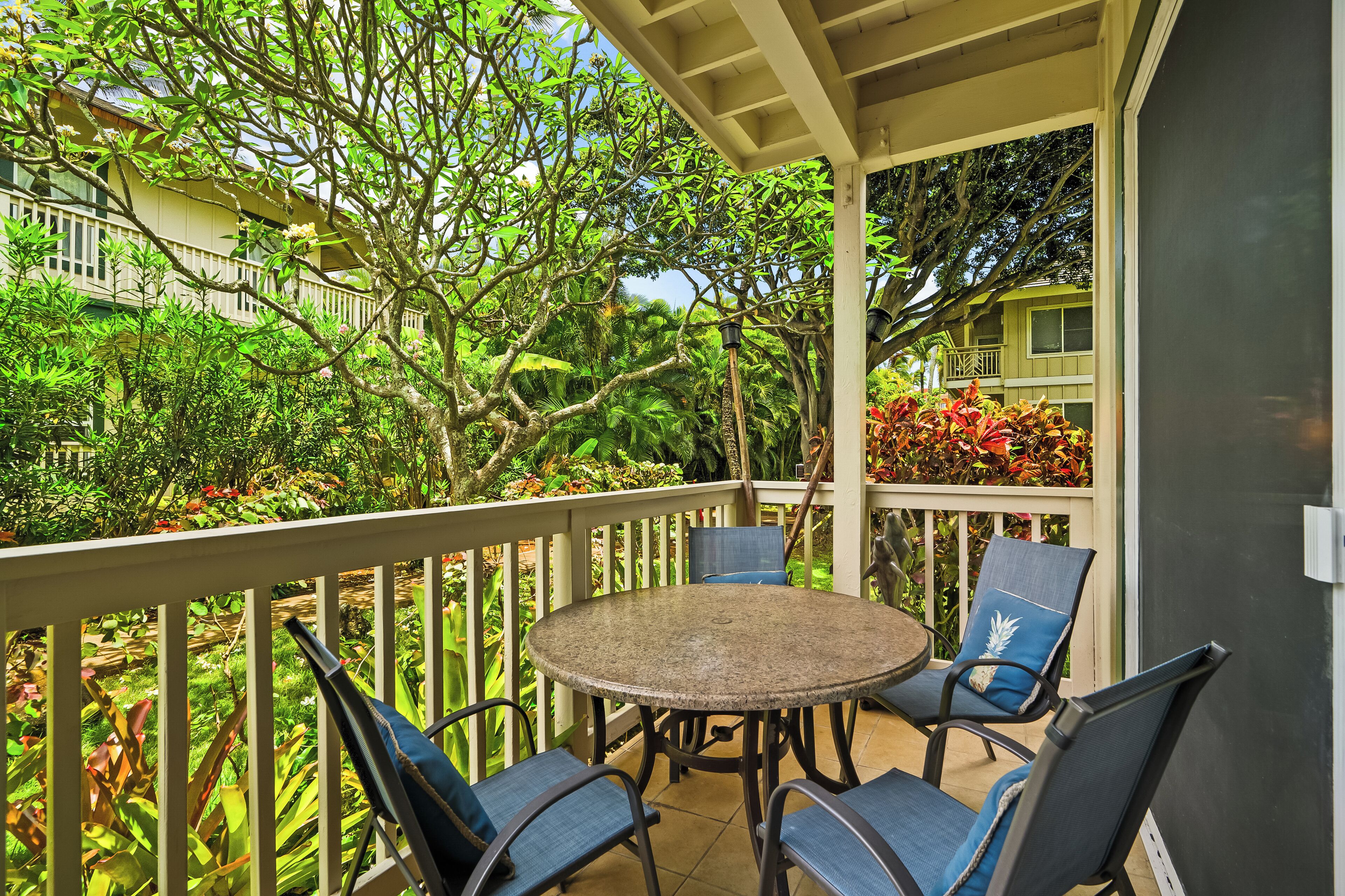 A covered outdoor seating area with a round table and four chairs, surrounded by a white railing, overlooking a lush garden and a building with balconies.