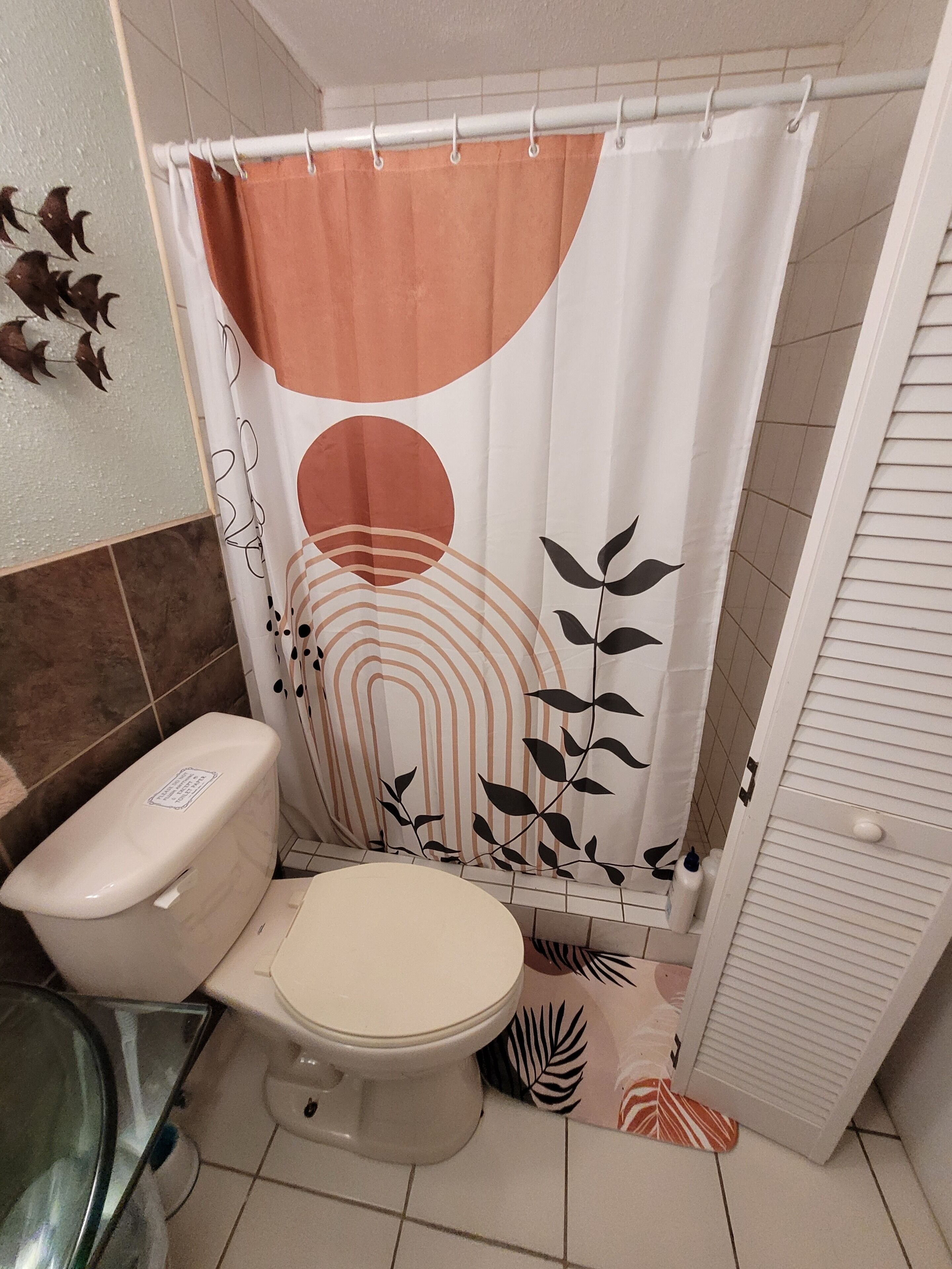 A bathroom with a toilet, a shower curtain with a leaf pattern, and a tiled floor.