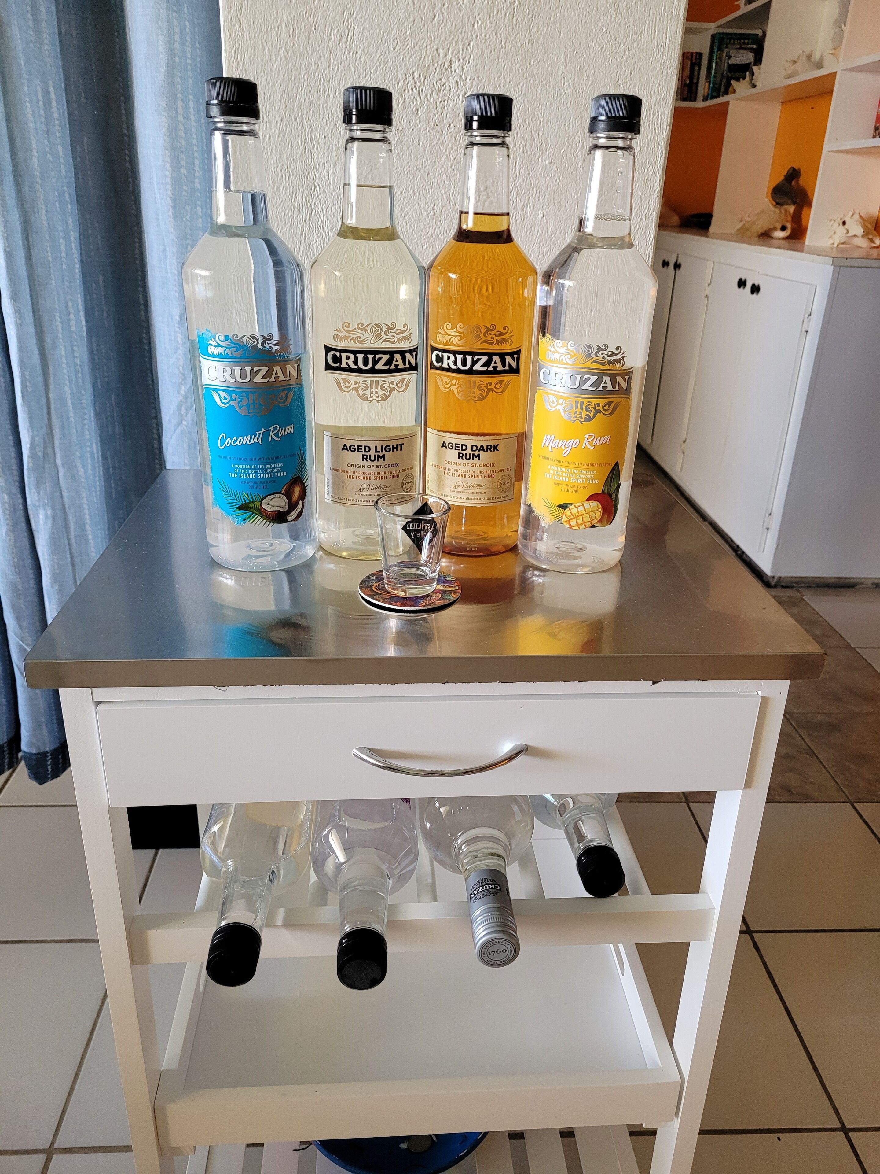 A white table with four bottles of Cruzan rum and a shot glass on top. Below, there are three empty bottles and a bottle with a black cap.