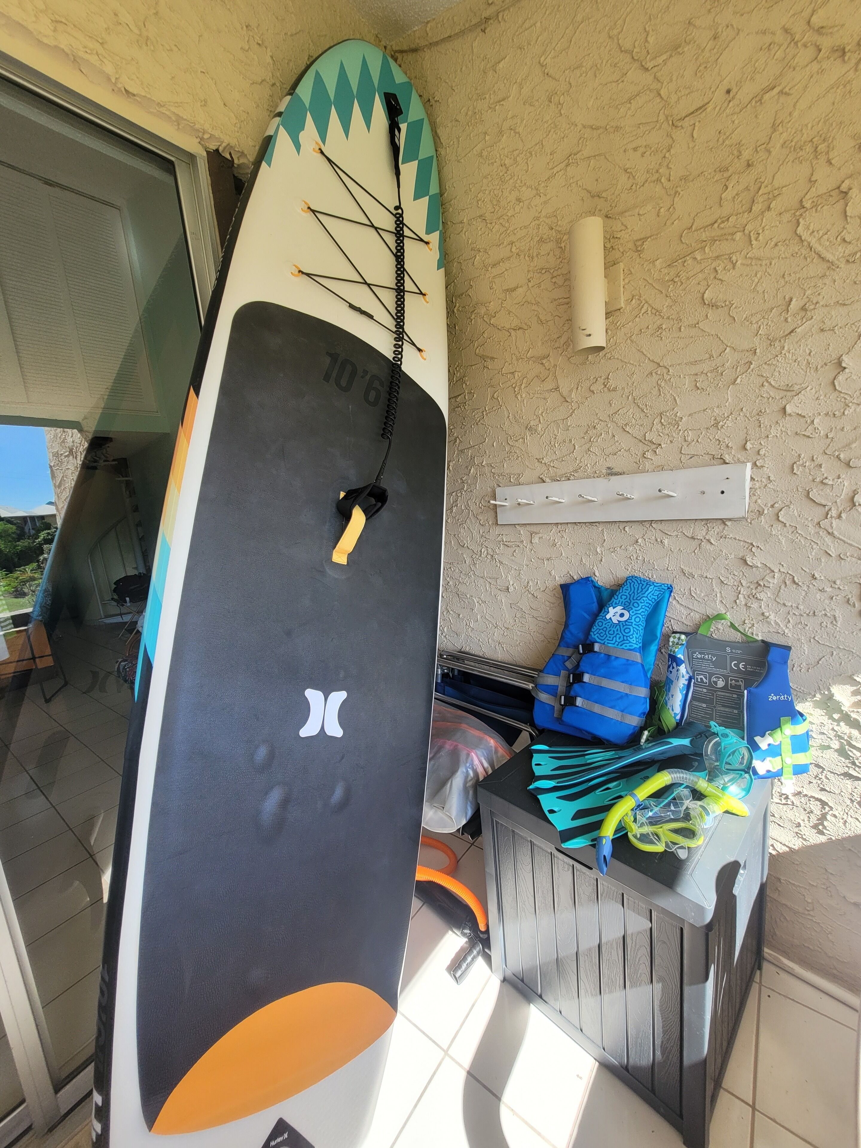 Una taula de surf recolzada contra una paret, un suport per a taules de surf i una taula amb diversos articles relacionats amb el surf.