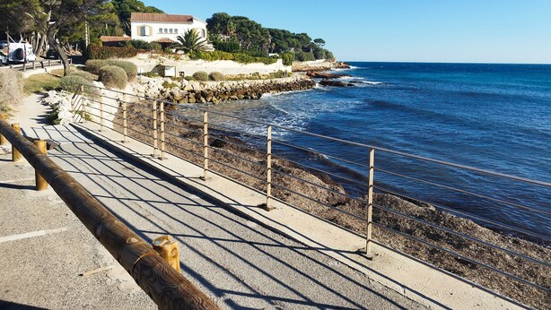 A coastal walkway with a stone wall, wooden railings, and a building with a tiled roof.