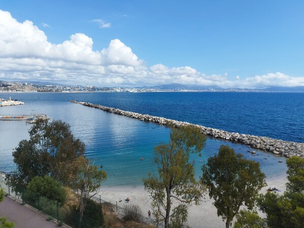 Vista costera con un rompeolas rocoso, agua azul cristalina y un horizonte urbano lejano.