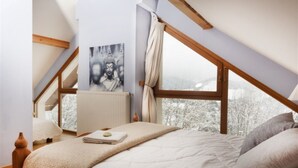 Ein Schlafzimmer mit schrägen Wänden, einem Bett mit einer beigen Decke, einem Heizkörper und einem Fenster mit Blick auf schneebedeckte Bäume.