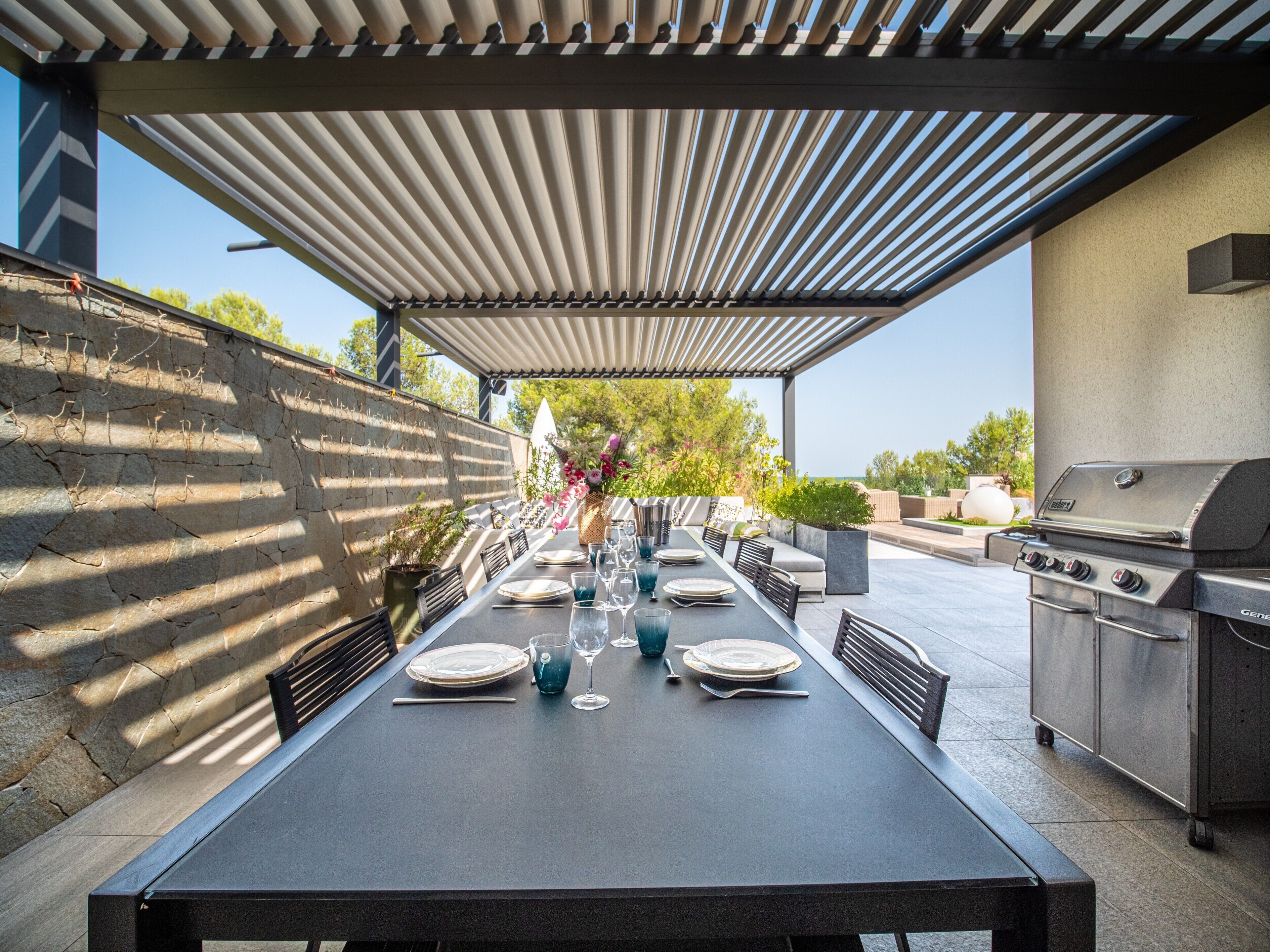 Une terrasse extérieure avec une table à manger pour six personnes, un barbecue et une pergola au-dessus.