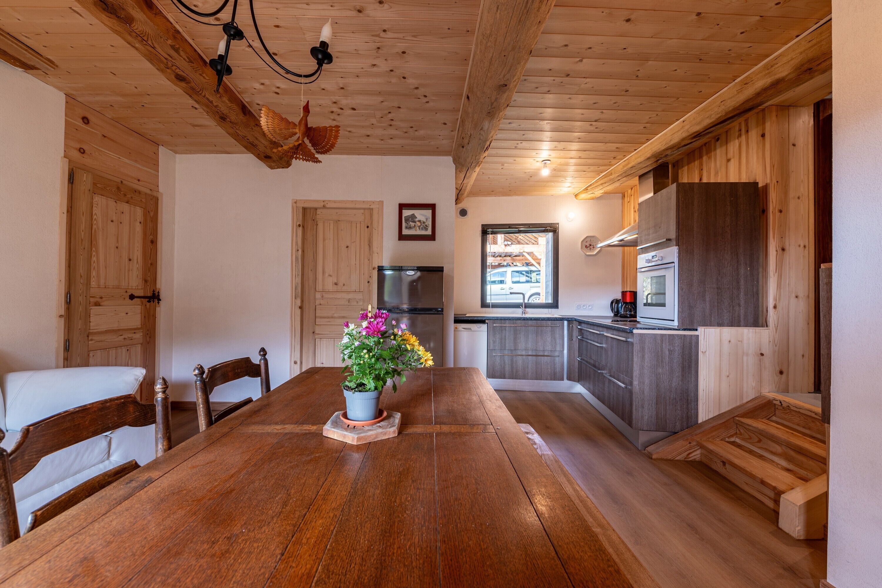 A wooden dining table with chairs, a potted plant, a wooden ceiling with exposed beams, a kitchen with modern appliances, and a view of the outside.