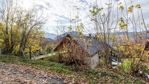 A rustic cabin with a sloped roof, surrounded by trees and a mountainous backdrop.