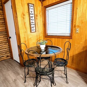Un interior de madera con una mesa redonda y cuatro sillas, una ventana con persianas, y un cartel que enumera productos agrícolas.