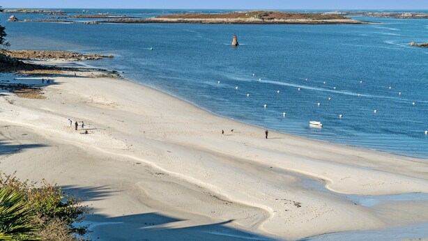 Une plage côtière avec un phare, une eau d’un bleu limpide et un rivage sablonneux.