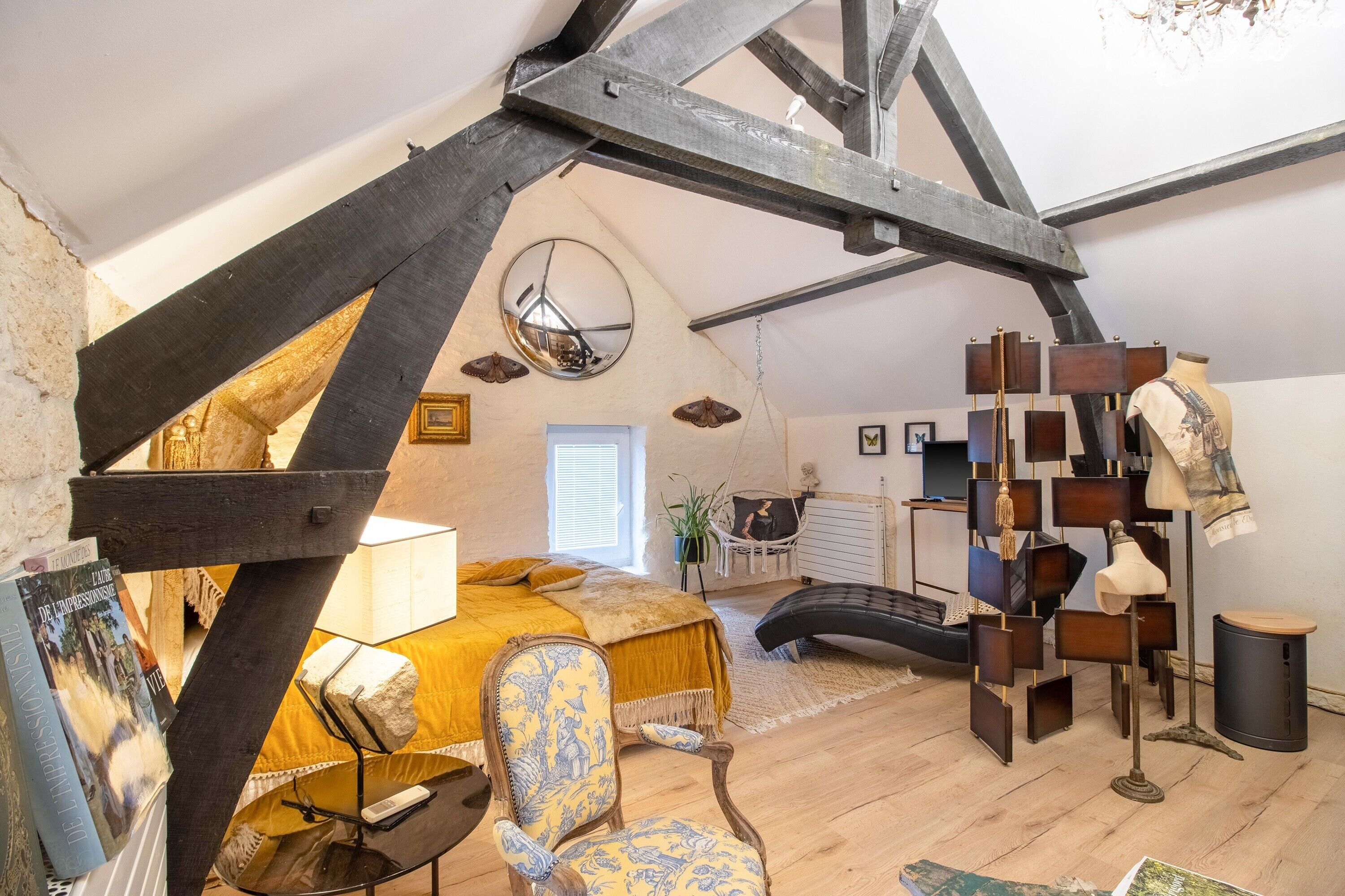 Ein Schlafzimmer mit schräg versiegelter Decke, einem Bett mit gelbem Kopfteil, einem Sessel mit gemustertem Kissen, einem Spiegel und einem außergewöhnlichen Holzregal.