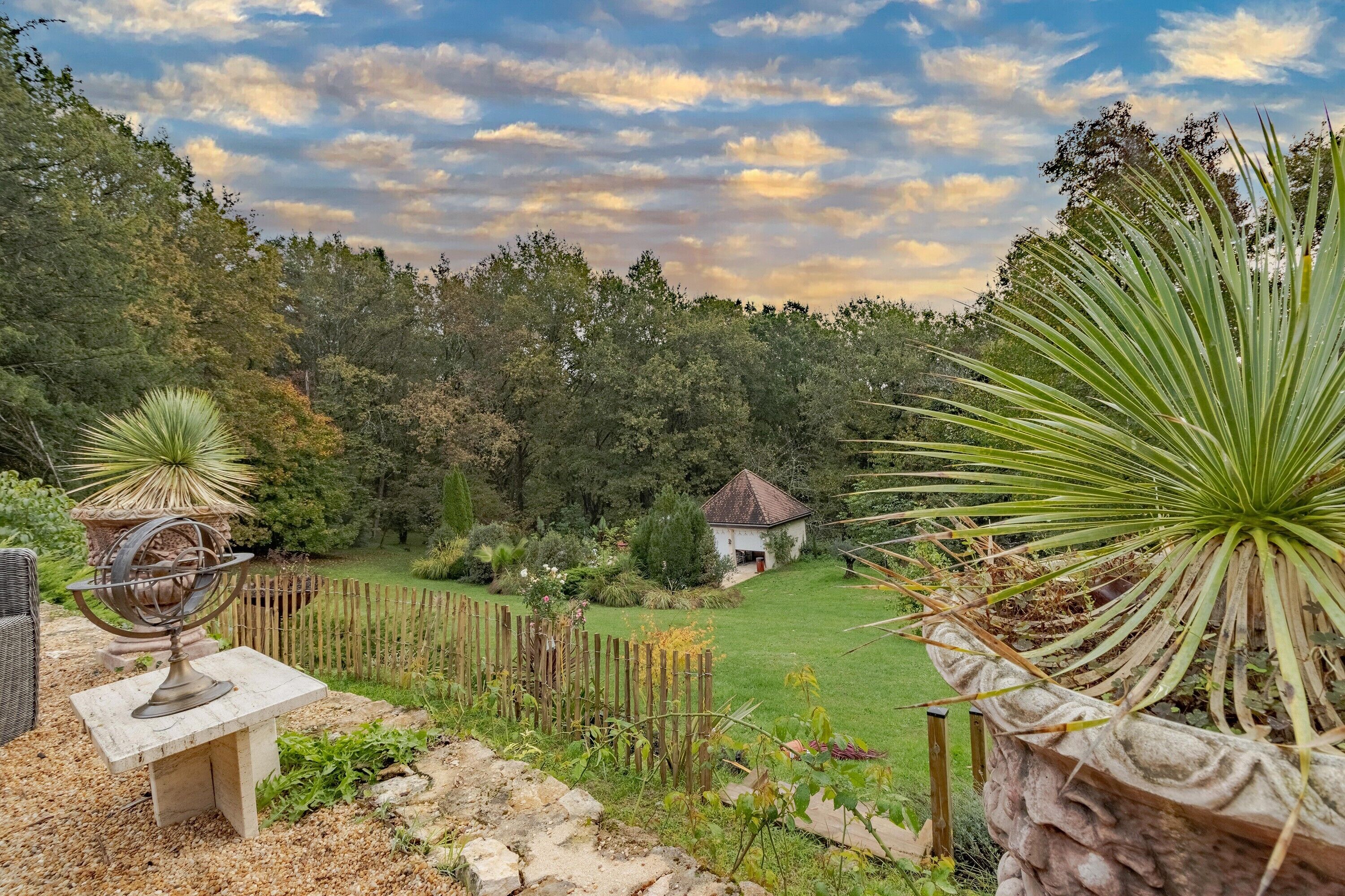 Un jardin avec un banc en pierre, une clôture en bois et une petite maison en arrière-plan.