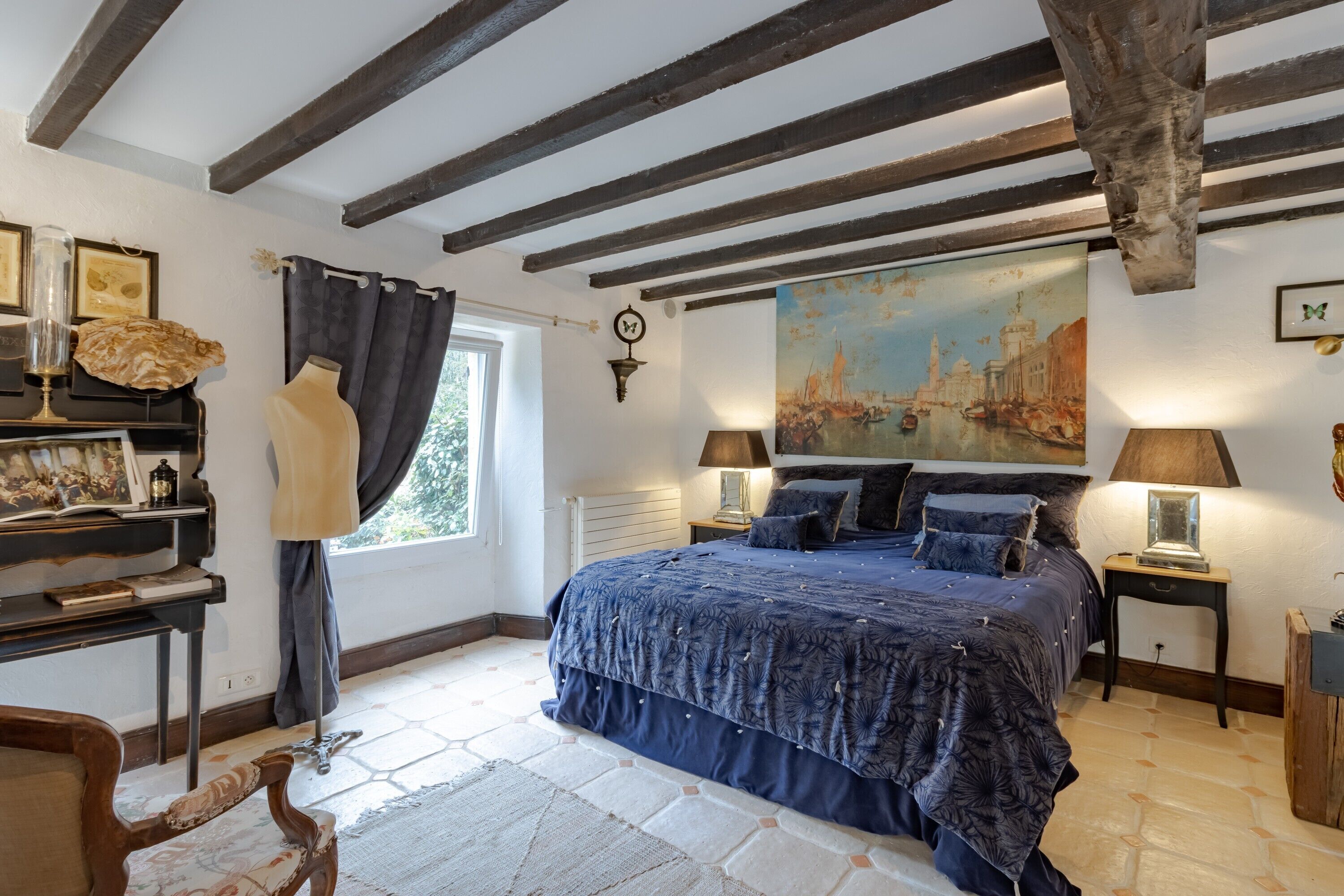 Ein Schlafzimmer mit einem großen Bett, einem Gemälde, einem Schreibtisch mit Lampe und einem Fenster mit Vorhängen.