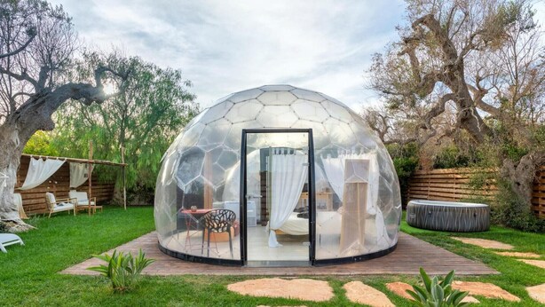 Une structure en forme de dôme géodésique dotée d’une porte en verre, d’un lit à l’intérieur et d’un espace patio aménagé avec des sièges et un jacuzzi.