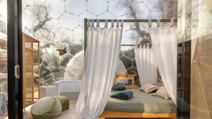Une chambre à coucher avec un lit à baldaquin, une table de chevet en bois, un canapé vert et une vue sur les arbres à travers une fenêtre hexagonale.